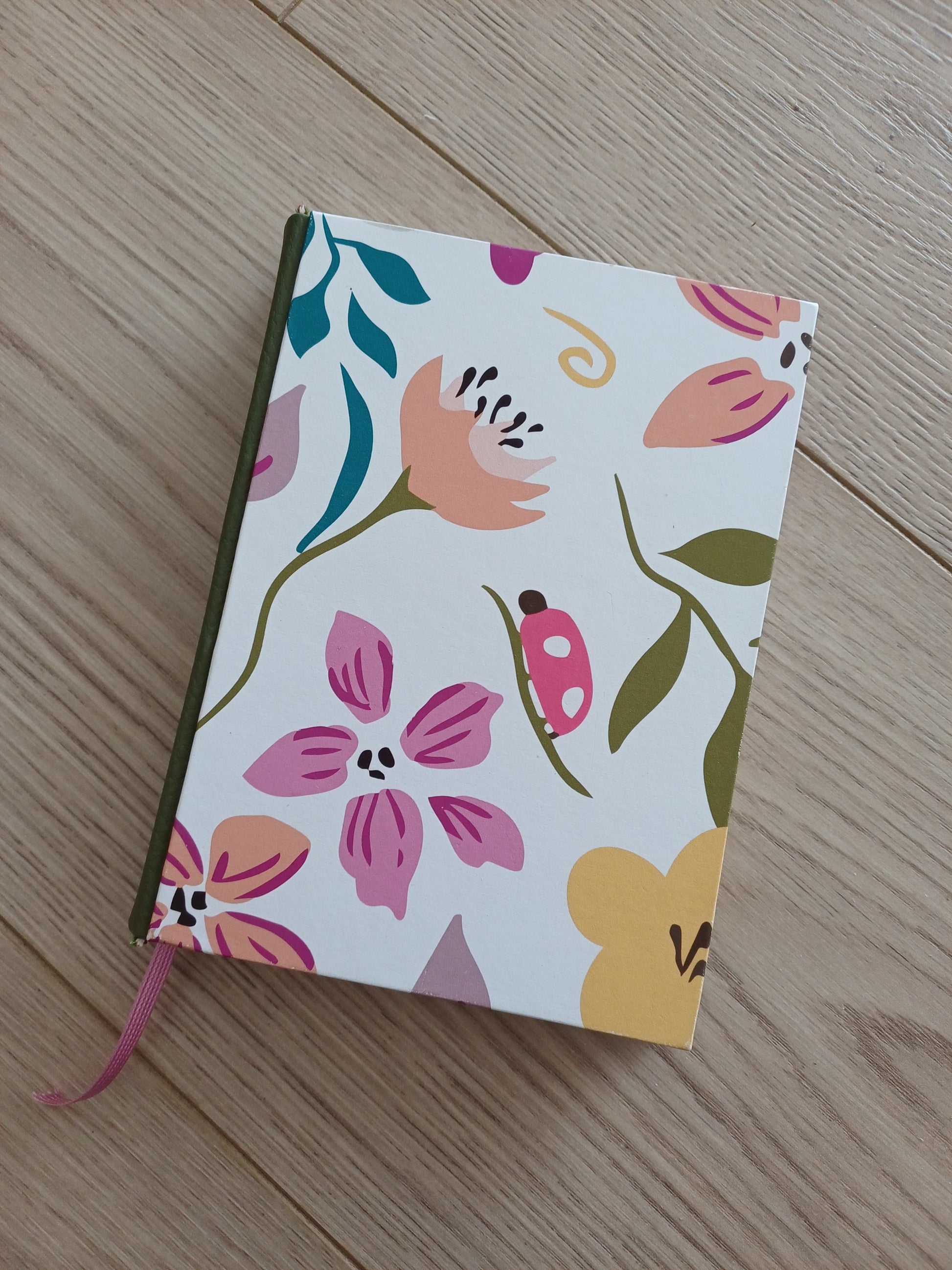 Carnet artisanal au dos en similicuir vert kaki et avec un signet rose. Sa couverture imprimée représente des dessin floraux avec des couleurs douces et joyeuses.