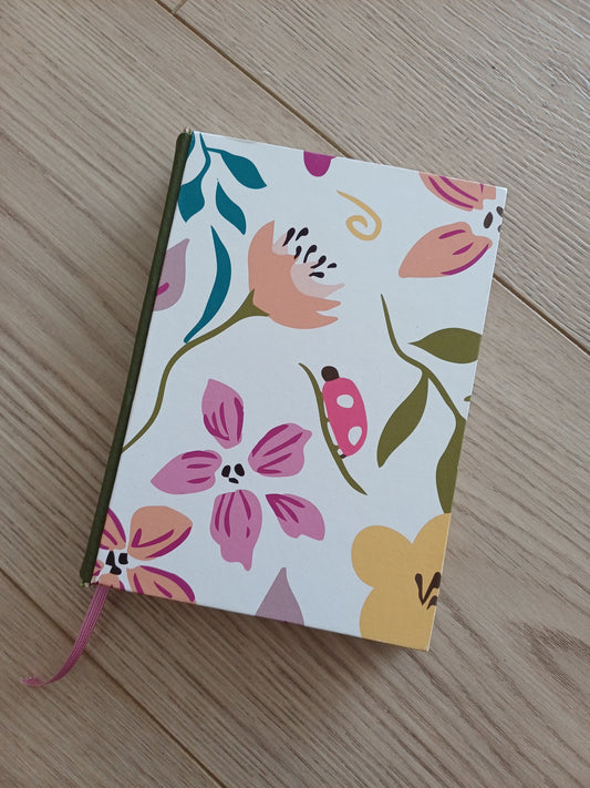 Carnet artisanal au dos en similicuir vert kaki et avec un signet rose. Sa couverture imprimée représente des dessin floraux avec des couleurs douces et joyeuses.