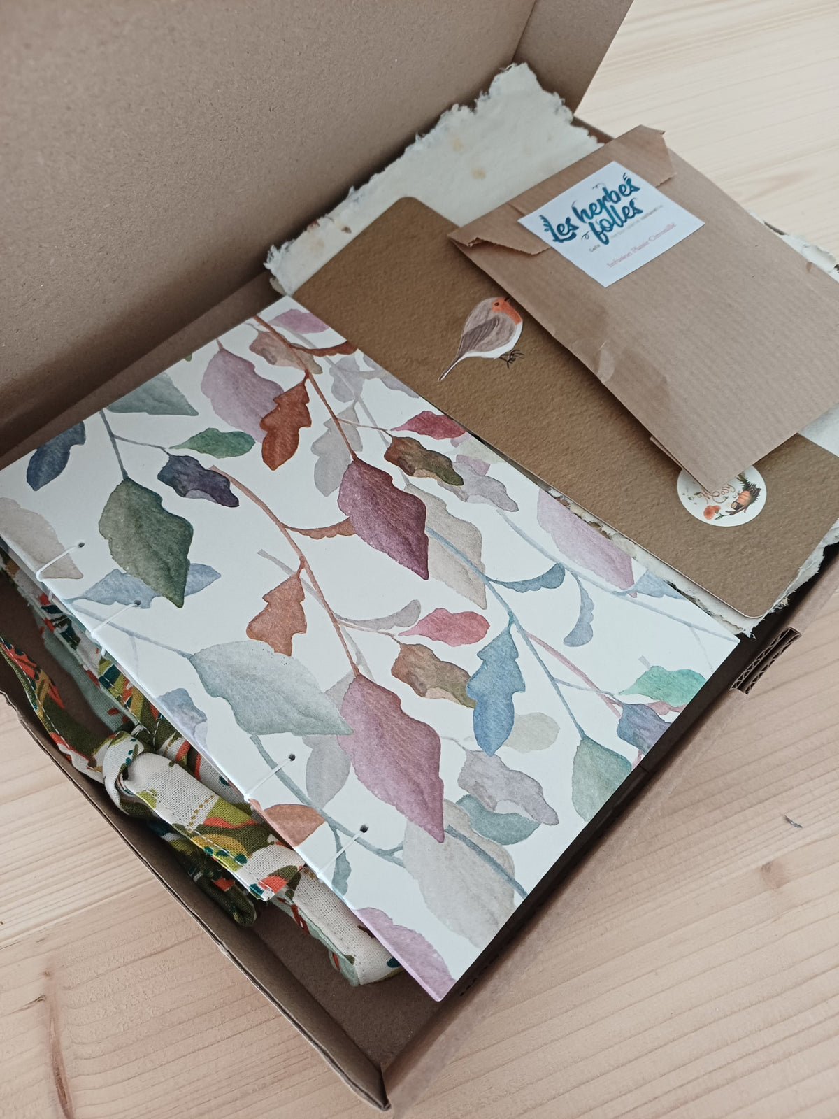 Un carnet artisanal posé dans une boite en carton, à côté de feuilles de papier recyclé, un marque page avec un dessin d'oiseau et un petit sachet en tissu beige avec une étiquette qui indique "Les herbes folles". En dessous on devine un objet en tissu avec des motifs floraux.
