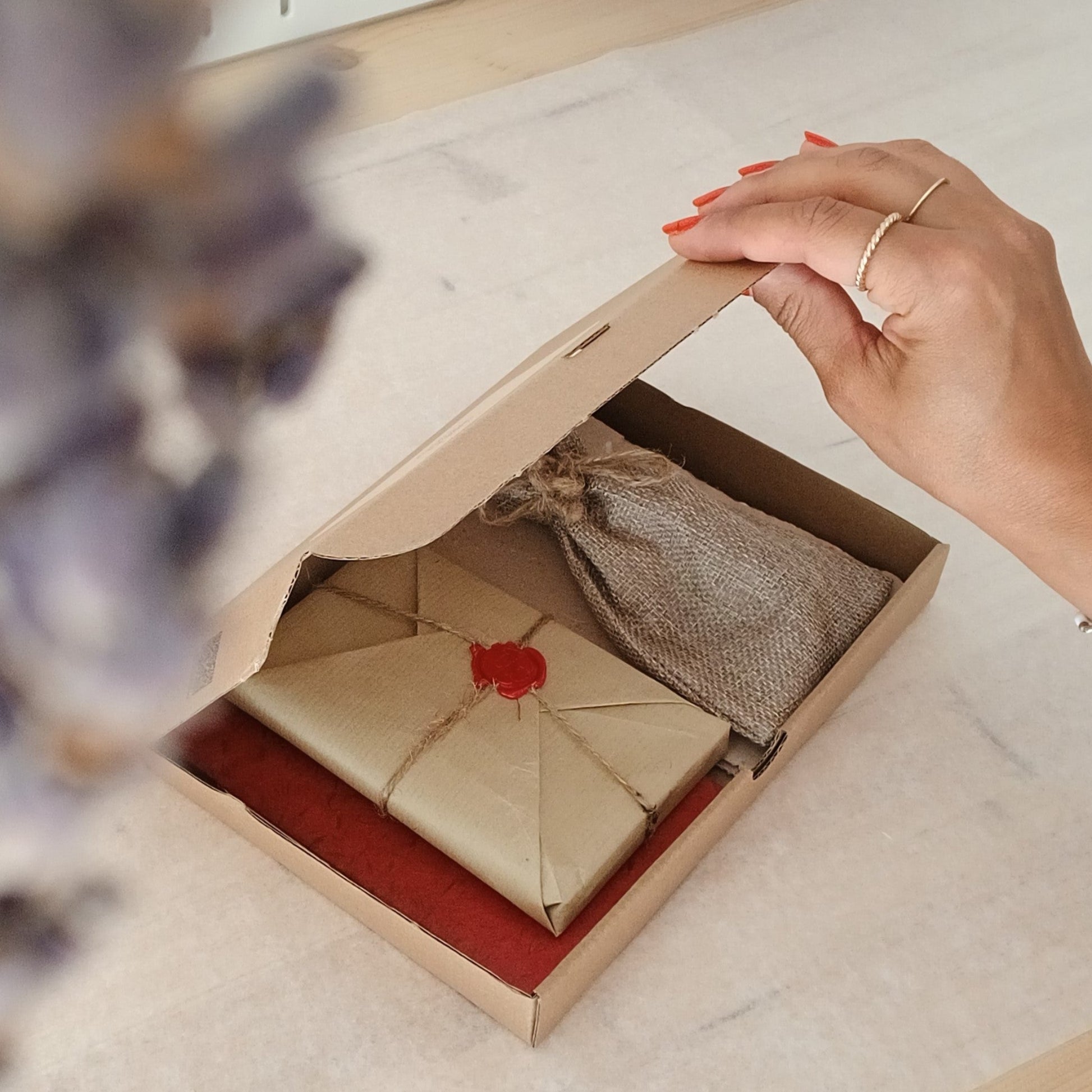 Une main ouvre légèrement une boite en carton contenant un paquet cadeau au style vintage et un pochon en jute.