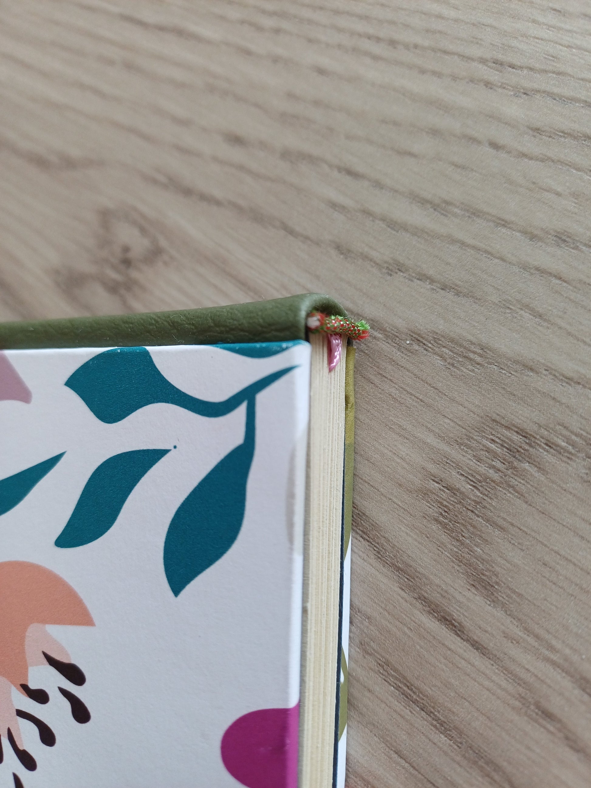 Tranche d'un carnet artisanal au dos en similicuir vert et à la couverture imprimée de motifs floraux colorés. On voit la tranche fil rouge et verte du carnet ainsi que le début du signet rose.
