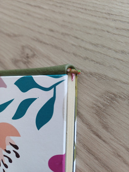 Tranche d'un carnet artisanal au dos en similicuir vert et à la couverture imprimée de motifs floraux colorés. On voit la tranche fil rouge et verte du carnet ainsi que le début du signet rose.