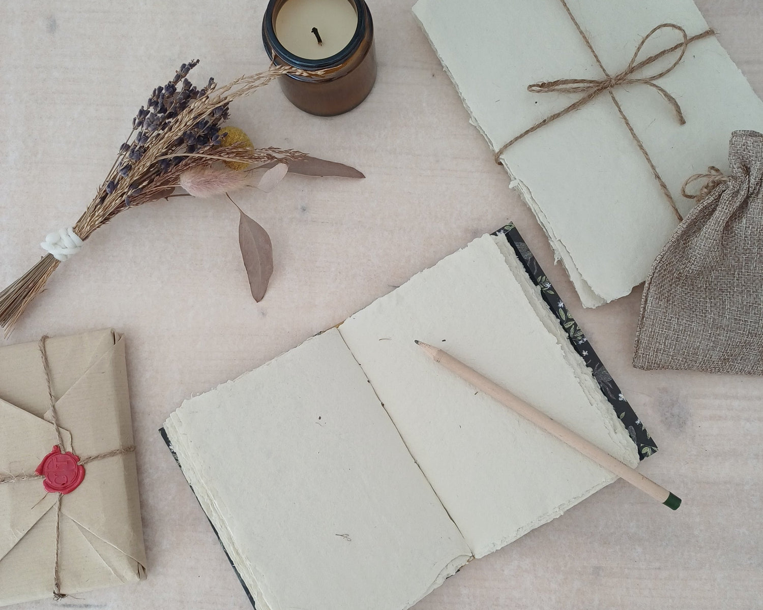 Carnet artisanal au papier blanc texturé recyclé sur lequel est posé un crayon de bois, à côté il y a un paquet de feuilles de papier recyclé emballé par du fil, une bougie éteinte, des fleurs séchées et un paquet cadeau au style vintage.