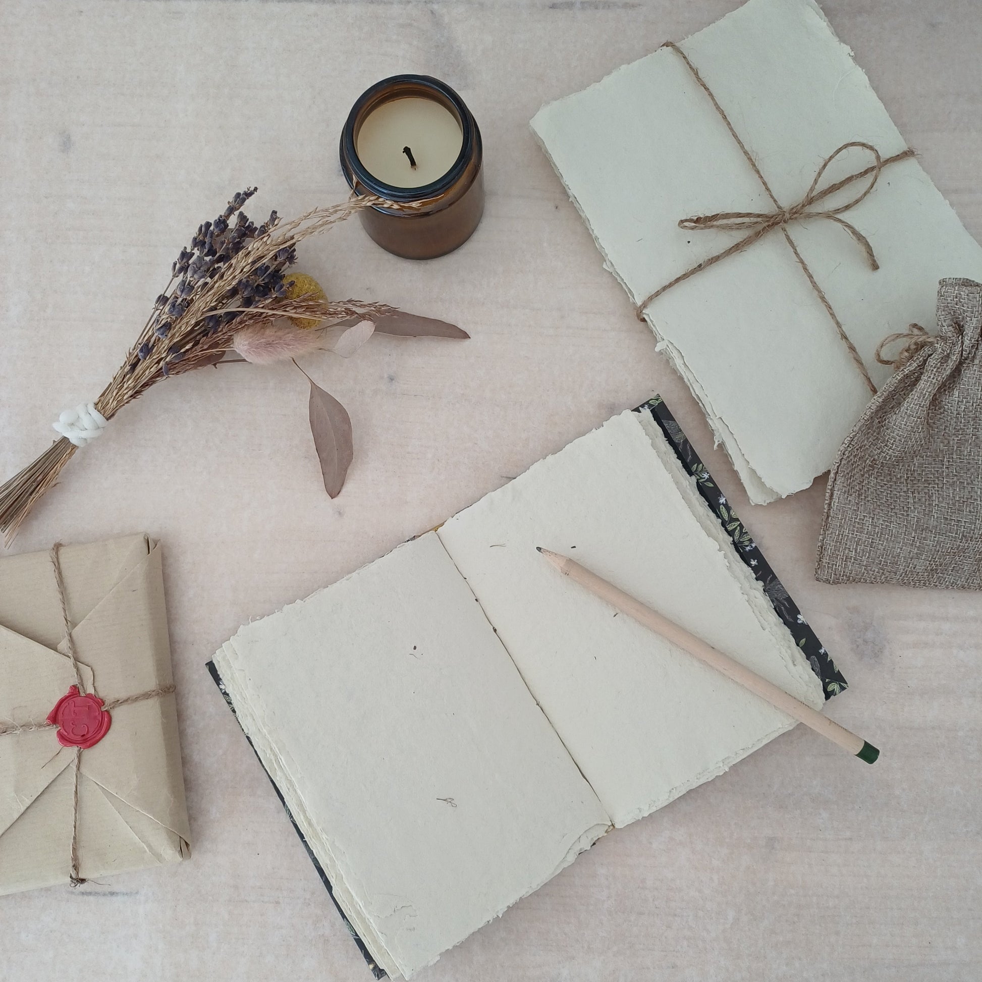 Carnet artisanal au papier blanc texturé recyclé sur lequel est posé un crayon de bois, à côté il y a un paquet de feuilles de papier recyclé emballé par du fil, une bougie éteinte, des fleurs séchées et un paquet cadeau au style vintage.
