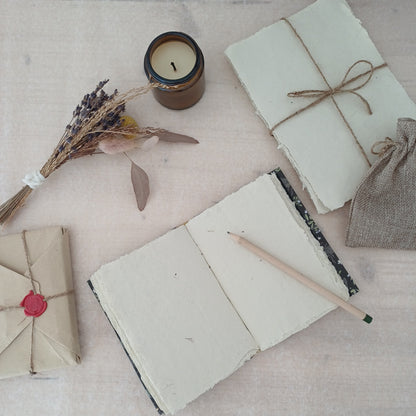 Carnet artisanal au papier blanc texturé recyclé sur lequel est posé un crayon de bois, à côté il y a un paquet de feuilles de papier recyclé emballé par du fil, une bougie éteinte, des fleurs séchées et un paquet cadeau au style vintage.