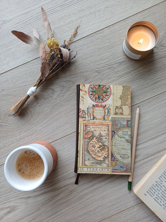 Carnet artisanal à la couverture en cartes du monde anciennes avec un crayon, un café, une bougie, des fleurs et un livre.