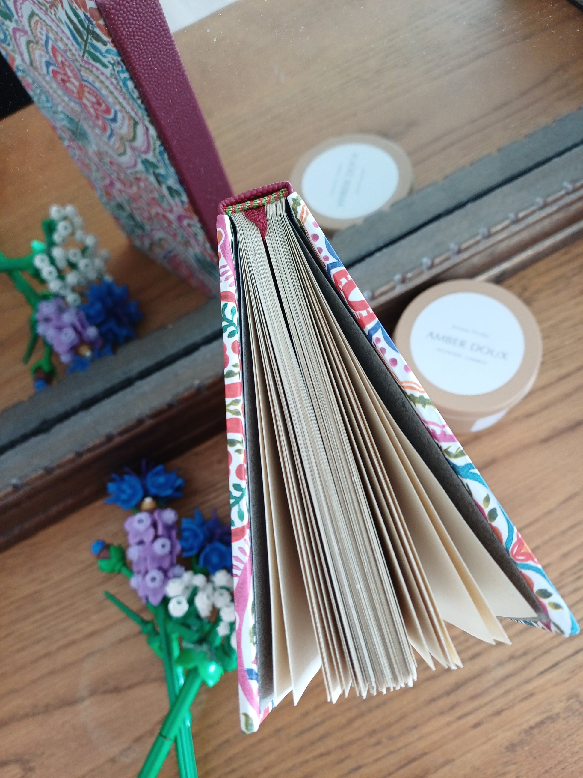 Carnet artisanal vue du dessus laissant voir la tranchefil et le signet. Il se reflète dans un miroir laissant voir sa couverture aux motifs de carreaux de faïence colorés, à côté de fleurs en legos et d'une bougie.