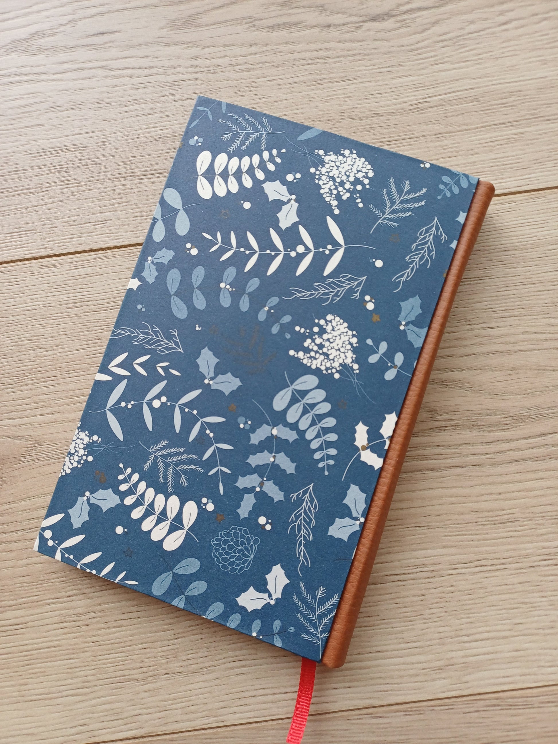 Carnet artisanal avec une couverture imprimée de motifs végétaux rappelant Noël sur fond bleu.