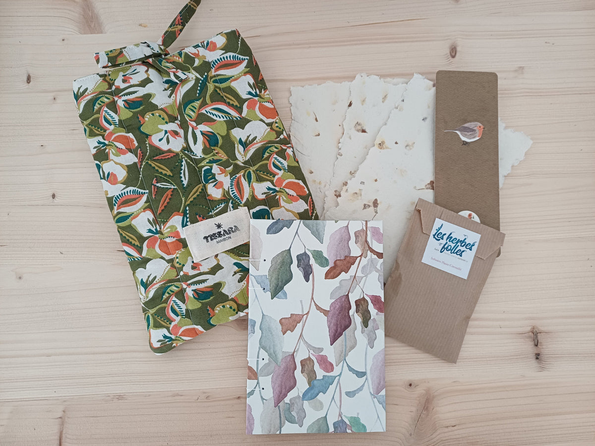 Un carnet artisanal posé sur du bois, à côté de 3 feuilles de papier recyclé, une pochette protège-livre avec des motifs floraux, un marque page avec un dessin d'oiseau et un petit sachet en tissu beige avec une étiquette qui indique "Les herbes folles".