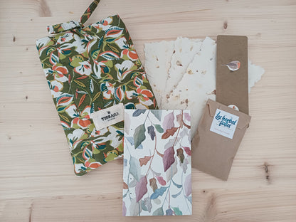Un carnet artisanal posé sur du bois, à côté de 3 feuilles de papier recyclé, une pochette protège-livre avec des motifs floraux, un marque page avec un dessin d'oiseau et un petit sachet en tissu beige avec une étiquette qui indique "Les herbes folles".