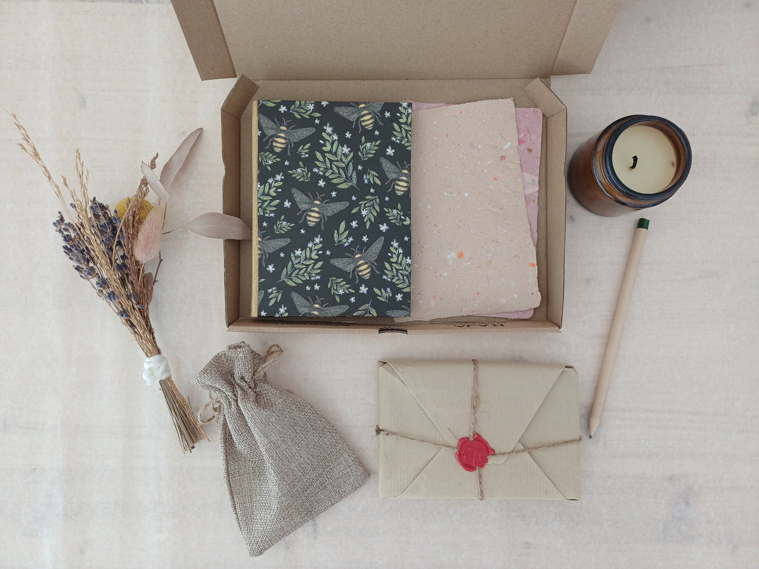 Une boite en carton contenant un carnet aux motifs végétaux et avec des abeilles, et du papier recyclé, à ôté d'une bougie éteinte, d'un petit bouquet de fleurs séchées, d'un crayon, d'un pochon et d'un paquet cadeau au style vintage.