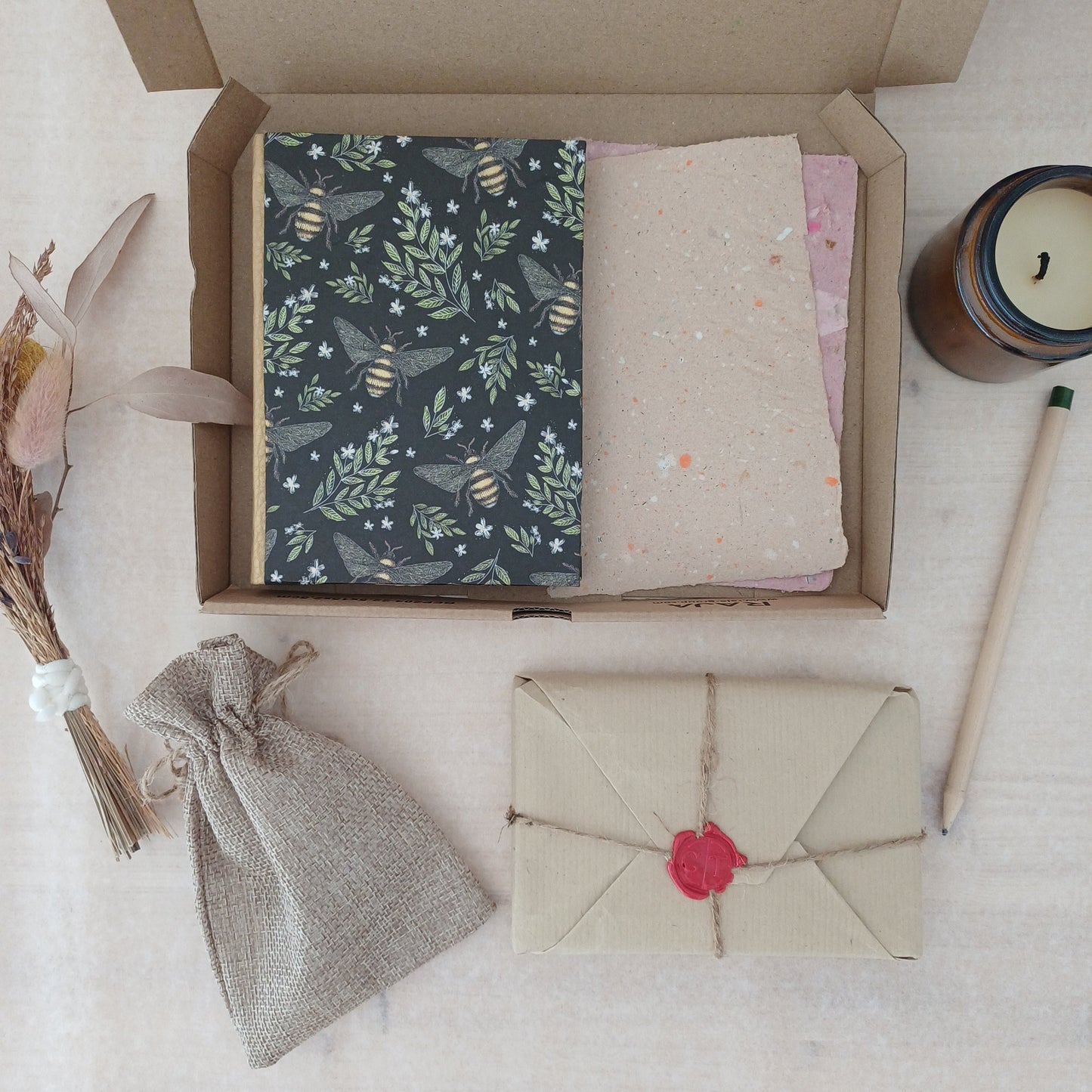 Une boite en carton contenant un carnet aux motifs végétaux et avec des abeilles, et du papier recyclé, à ôté d'une bougie éteinte, d'un petit bouquet de fleurs séchées, d'un crayon, d'un pochon et d'un paquet cadeau au style vintage.