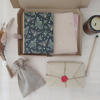 Une boite en carton contenant un carnet aux motifs végétaux et avec des abeilles, et du papier recyclé, à ôté d'une bougie éteinte, d'un petit bouquet de fleurs séchées, d'un crayon, d'un pochon et d'un paquet cadeau au style vintage.