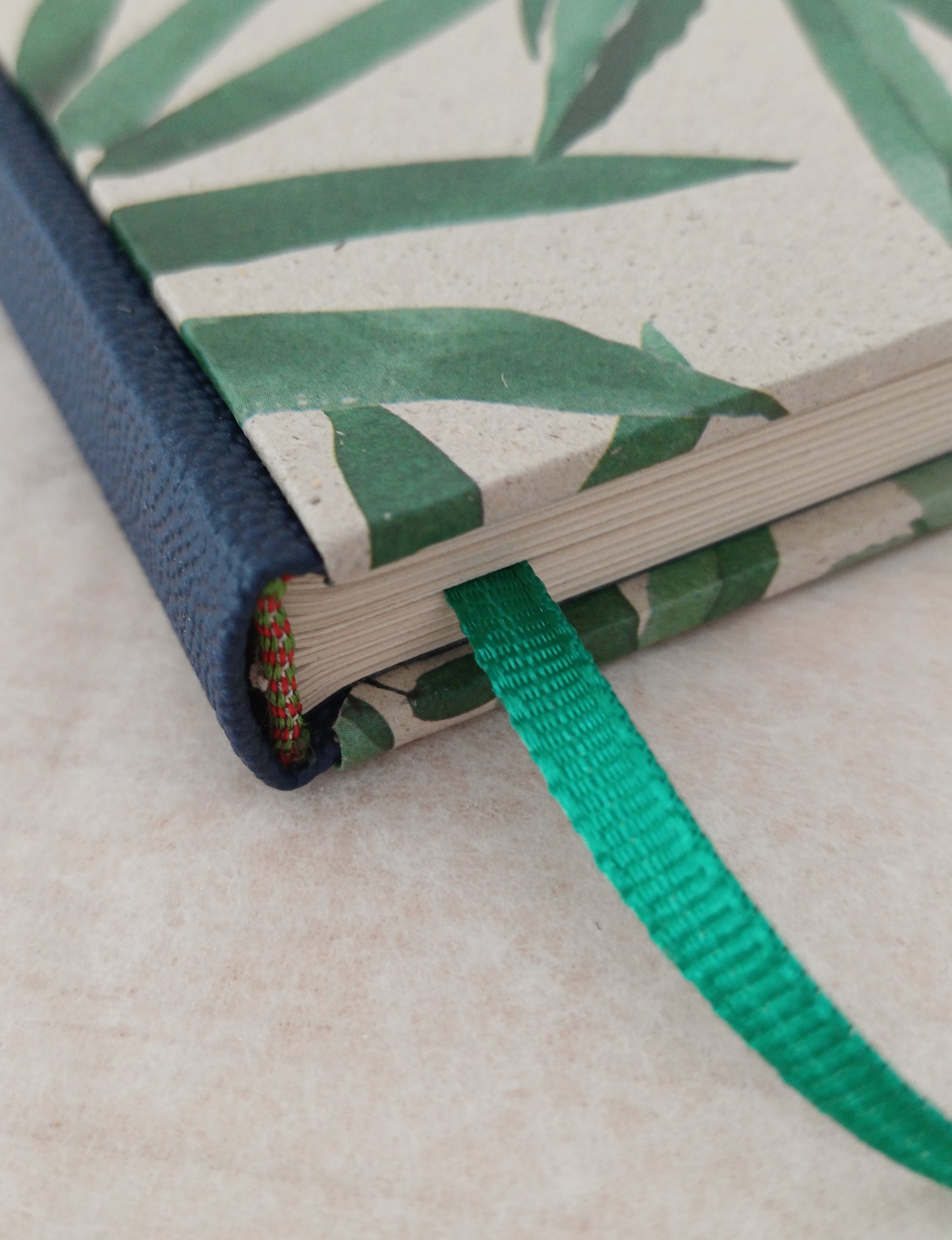 Détail d'un carnet artisanal avec une couverture aux figures végétales,  montrant une partie du dos en faux cuir bleu, la tranchefile et le signet vert.