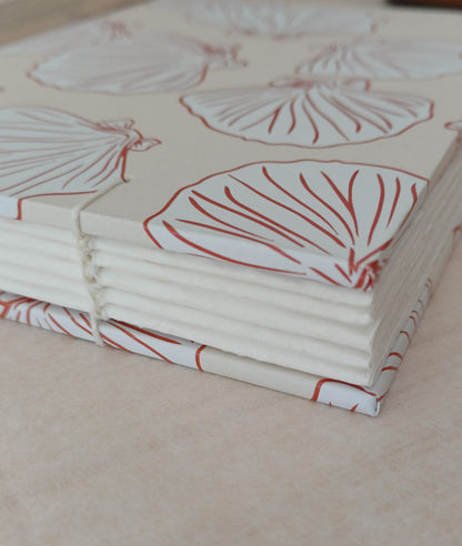 Détail d'un carnet artisanal relié en reliure copte visible. La couverture du carnet présente des motifs de coquillages de couleur beige et terracotta.
