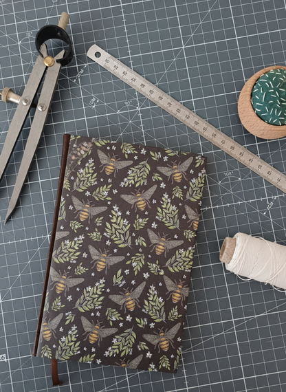 Carnet à la couverture imprimée avec des abeilles et des feuillages, entouré d'un compas, d'un réglet d'aiguilles et de fil.
