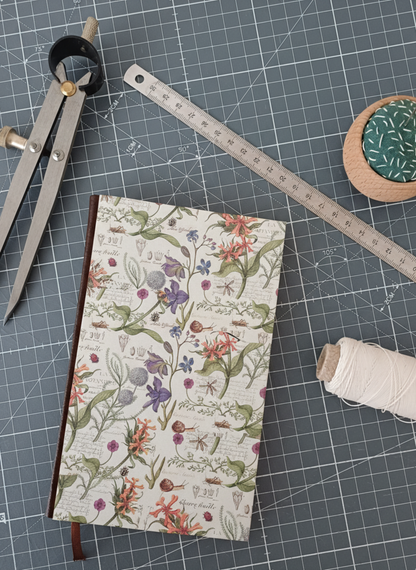 Carnet à la couverture imprimée de motifs de botanique entouré d'un compas, d'un réglet d'aiguilles et de fil.