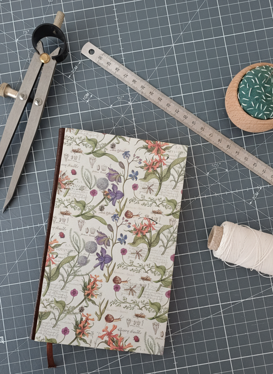Carnet à la couverture imprimée de motifs de botanique entouré d'un compas, d'un réglet d'aiguilles et de fil.