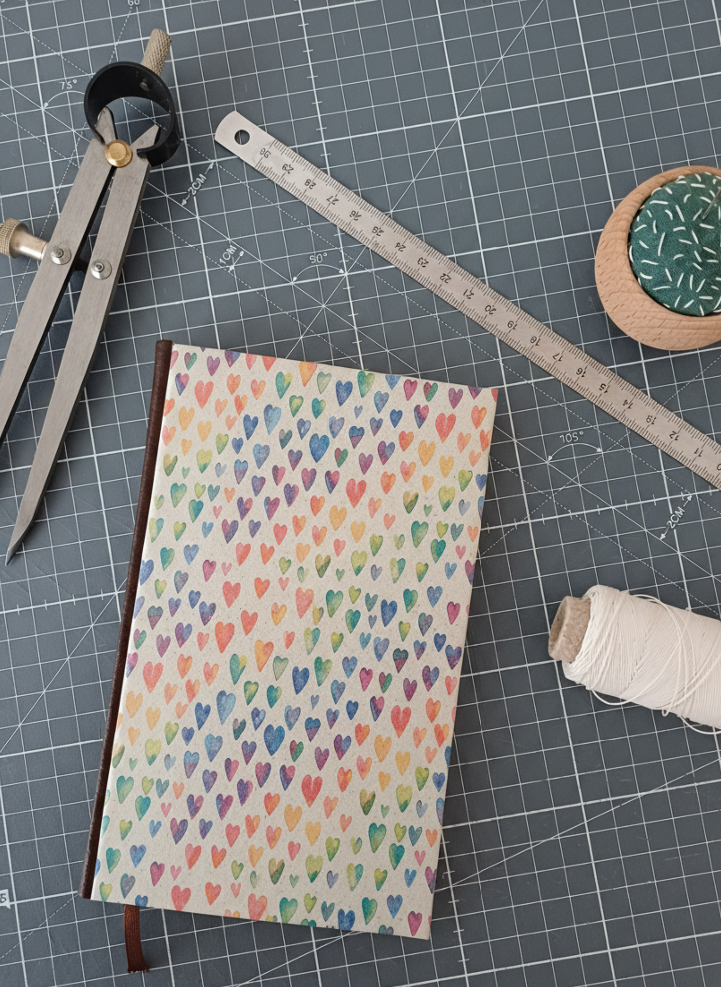 Carnet à la couverture imprimée de cœurs aux couleurs de l'arc-en-ciel, entouré d'un compas, d'un réglet d'aiguilles et de fil.