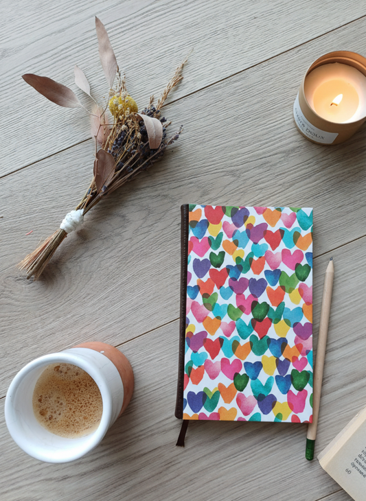 Carnet à la couverture imprimée avec des cœurs colorés, entouré de fleurs, d'un café, d'une bougie, d'un livre et d'un crayon.