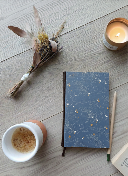 Carnet à la couverture imprimée de constellations, entouré de fleurs, d'un café, d'une bougie, d'un livre et d'un crayon.