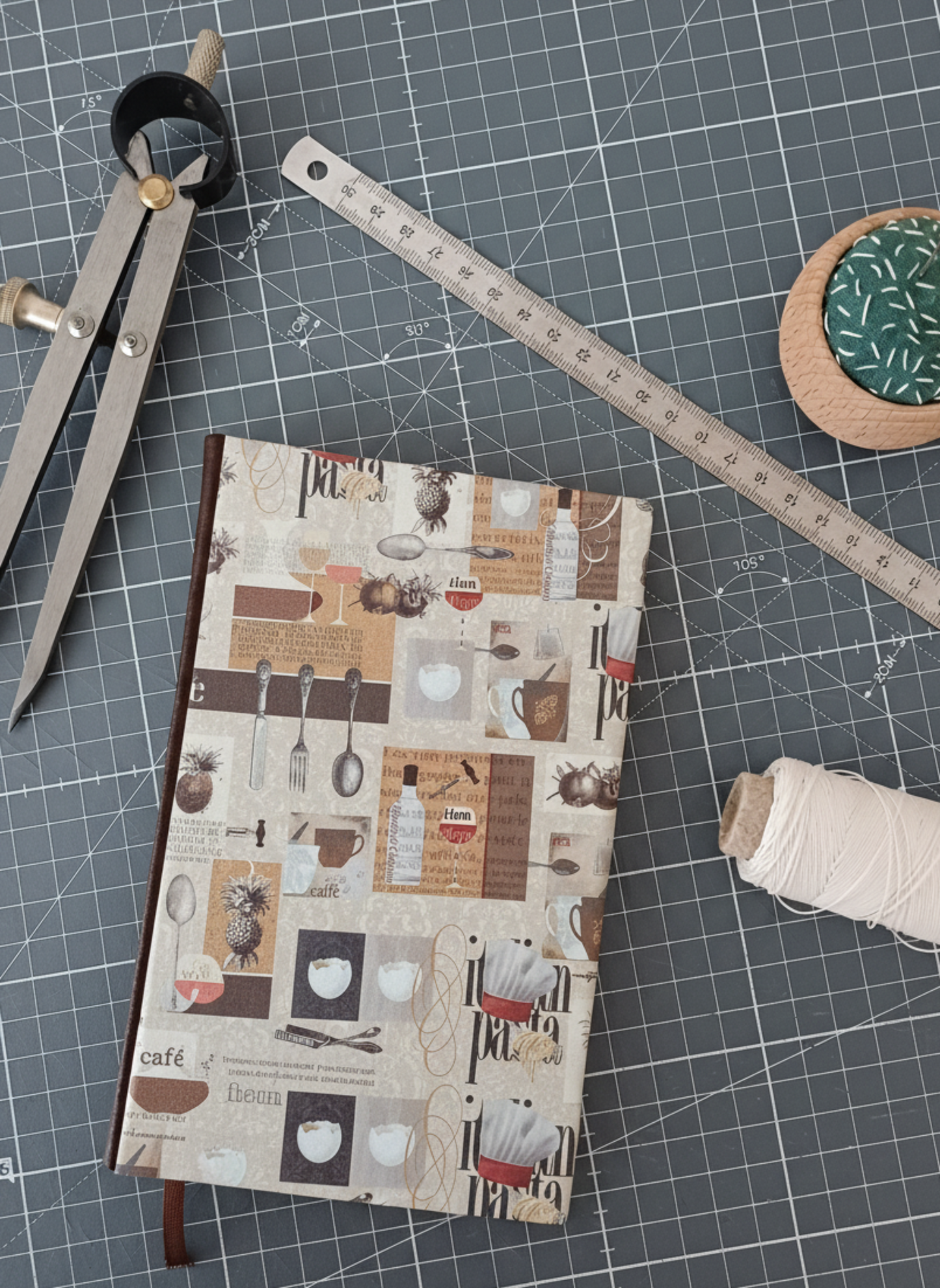 Carnet à la couverture imprimée de dessins d'ustensiles évoquant la cuisine italienne, entouré d'un compas, d'un réglet d'aiguilles et de fil.