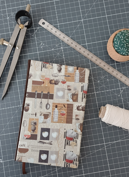 Carnet à la couverture imprimée de dessins d'ustensiles évoquant la cuisine italienne, entouré d'un compas, d'un réglet d'aiguilles et de fil.