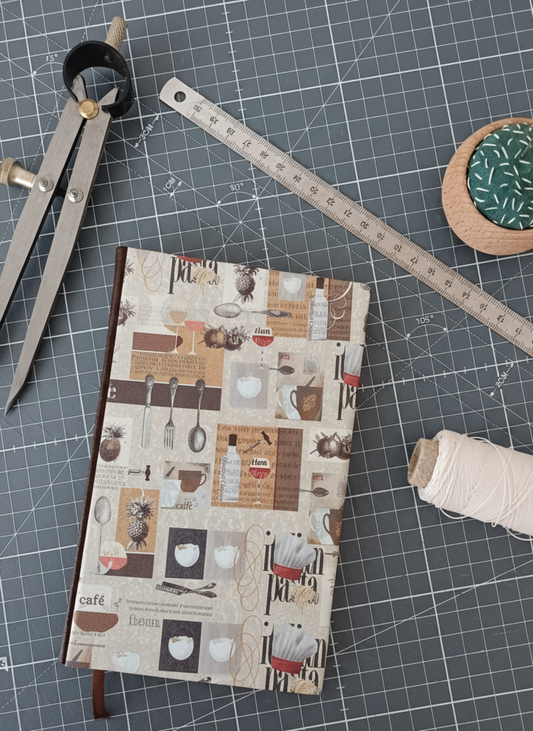 Carnet à la couverture imprimée de dessins d'ustensiles évoquant la cuisine italienne, entouré d'un compas, d'un réglet d'aiguilles et de fil.