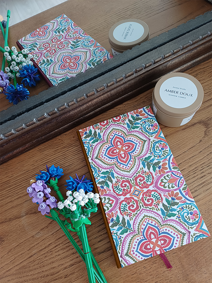 Carnet fait main avec une couverture imprimée aux motifs de carreaux de faïence colorée et un dos marron clair, à côté de fleurs en legos et d'une bougie, le tout se reflétant dans un miroir.