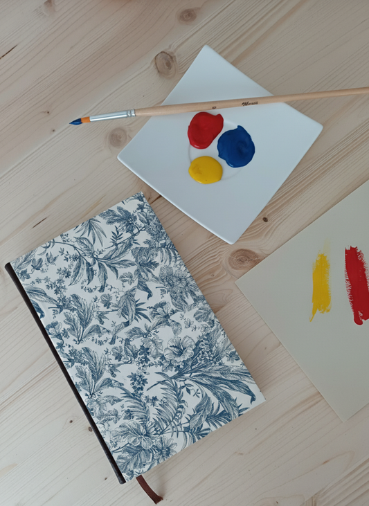 Carnet à la couverture imprimé de dessins fleuris bleus à côté d'une palette de peinture et un pinceau.
