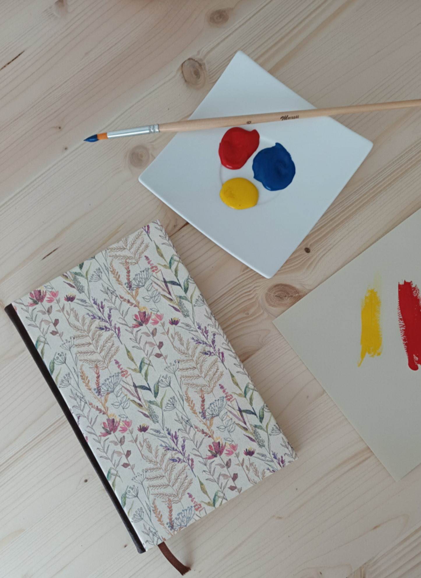 Carnet à la couverture imprimé d'un motif de fleurs sauvages à côté d'une palette de peinture et un pinceau.