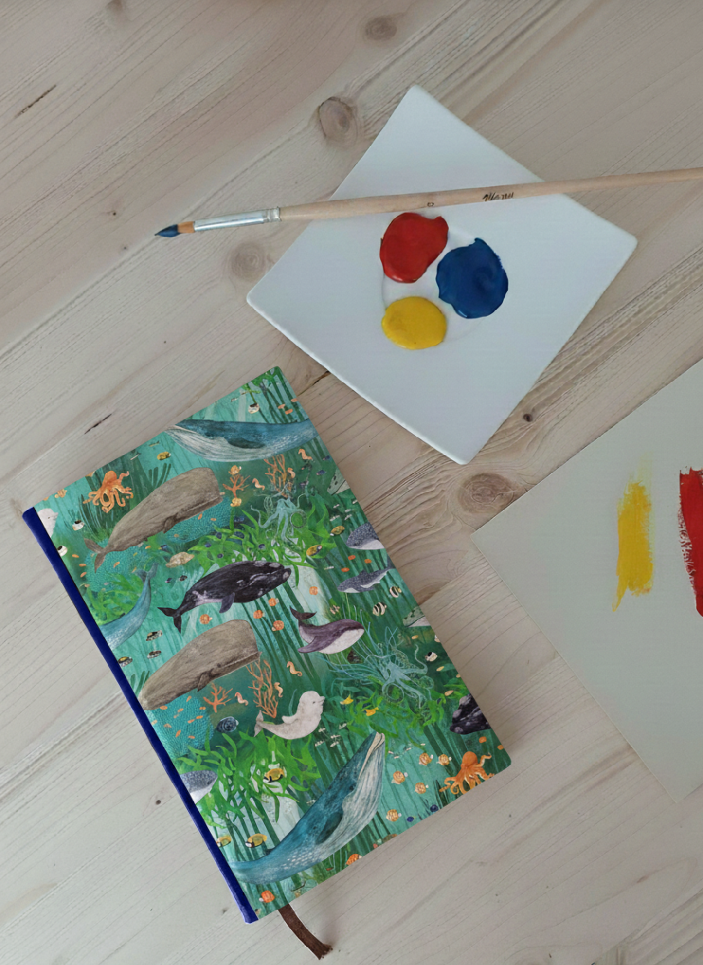 Carnet artisanal au dos en faux cuir bleu avec une couverture imprimée d'animaux marins et d'algues et autres plantes marines à côté d'une palette avec de la peinture et un pinceau.