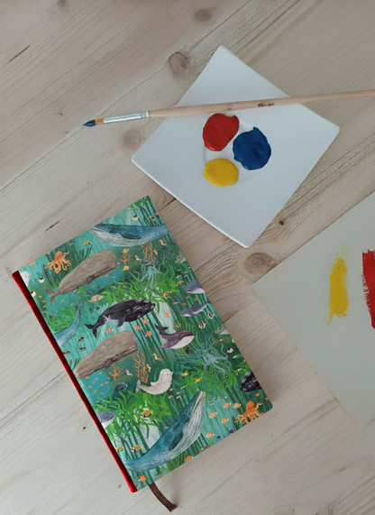 Carnet artisanal au dos en faux cuir rouge avec une couverture imprimée d'animaux marins et d'algues et autres plantes marines à côté d'une palette avec de la peinture et un pinceau.