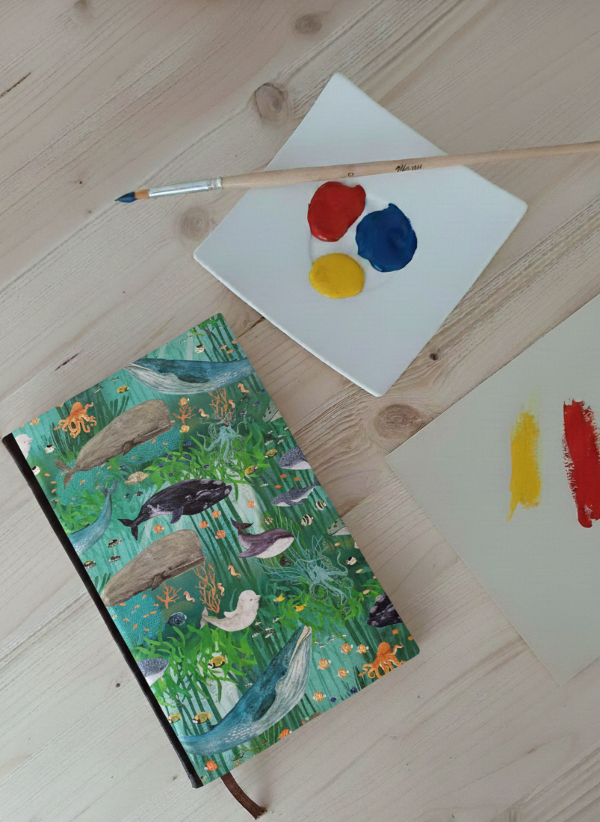 Carnet à la couverture imprimé d'un dessin de fons marins à côté d'une palette de peinture et un pinceau.
