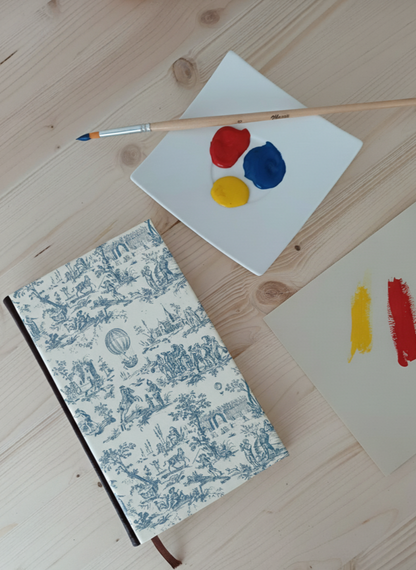 Carnet à la couverture imprimé de motifs vintage de toile de jouy à côté d'une palette de peinture et un pinceau.