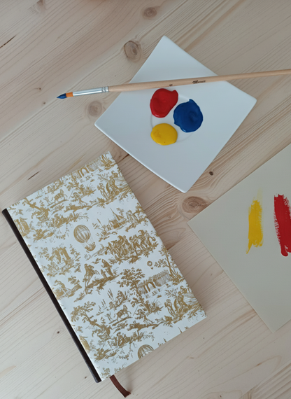 Carnet à la couverture imprimé de dessins vintage de toile de jouy à côté d'une palette de peinture et un pinceau.