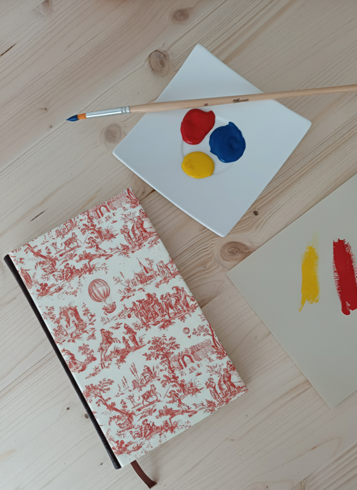 Carnet à la couverture imprimé de dessins vintage de toile de jouy  à côté d'une palette de peinture et un pinceau.