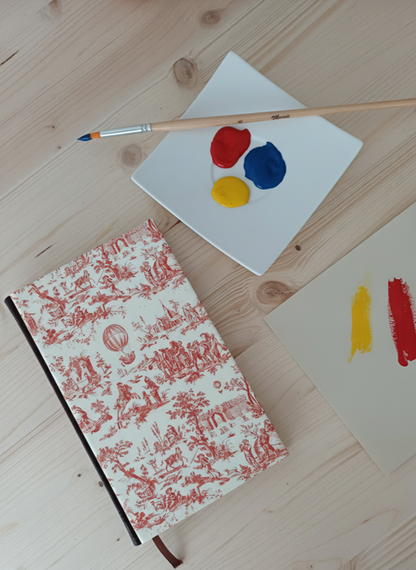 Carnet à la couverture imprimé de dessins vintage de toile de jouy  à côté d'une palette de peinture et un pinceau.