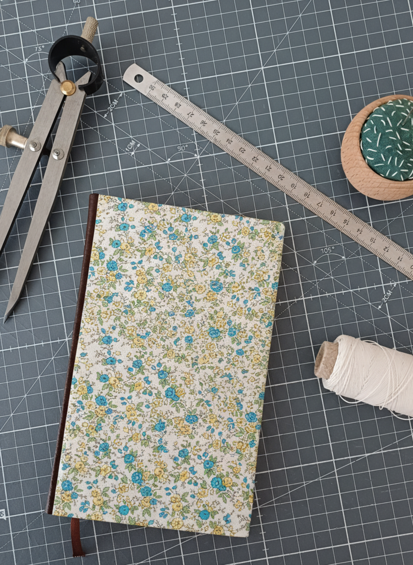 Carnet à la couverture imprimée de motifs fleuris style Liberty entouré d'un compas, d'un réglet d'aiguilles et de fil.