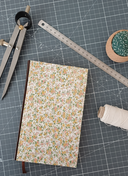 Carnet à la couverture imprimée de motifs fleuris style Liberty corail et jaune, entouré d'un compas, d'un réglet d'aiguilles et de fil.