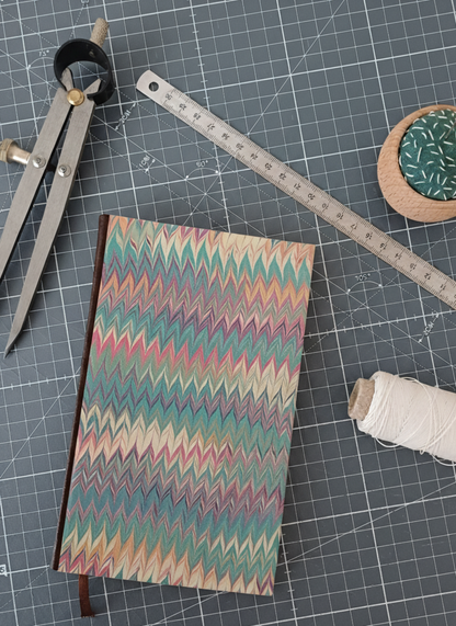 Carnet à la couverture imprimée de motifs marbrés en chevrons rouges et verts, entouré d'un compas, d'un réglet d'aiguilles et de fil.
