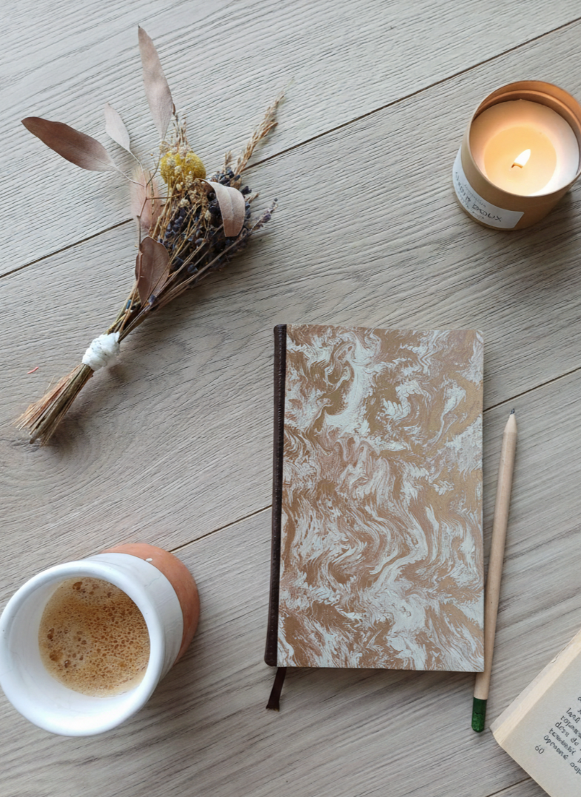 Carnet à la couverture imprimée avec des motifs marbrés dorés, entouré de fleurs, d'un café, d'une bougie, d'un livre et d'un crayon.