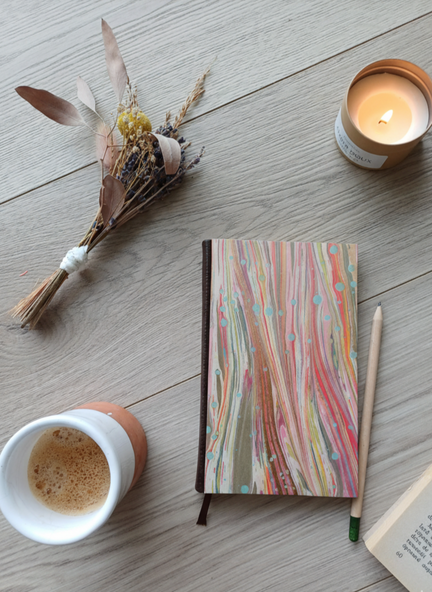 Carnet à la couverture imprimée avec des motifs marbrés roses et dorés, entouré de fleurs, d'un café, d'une bougie, d'un livre et d'un crayon.