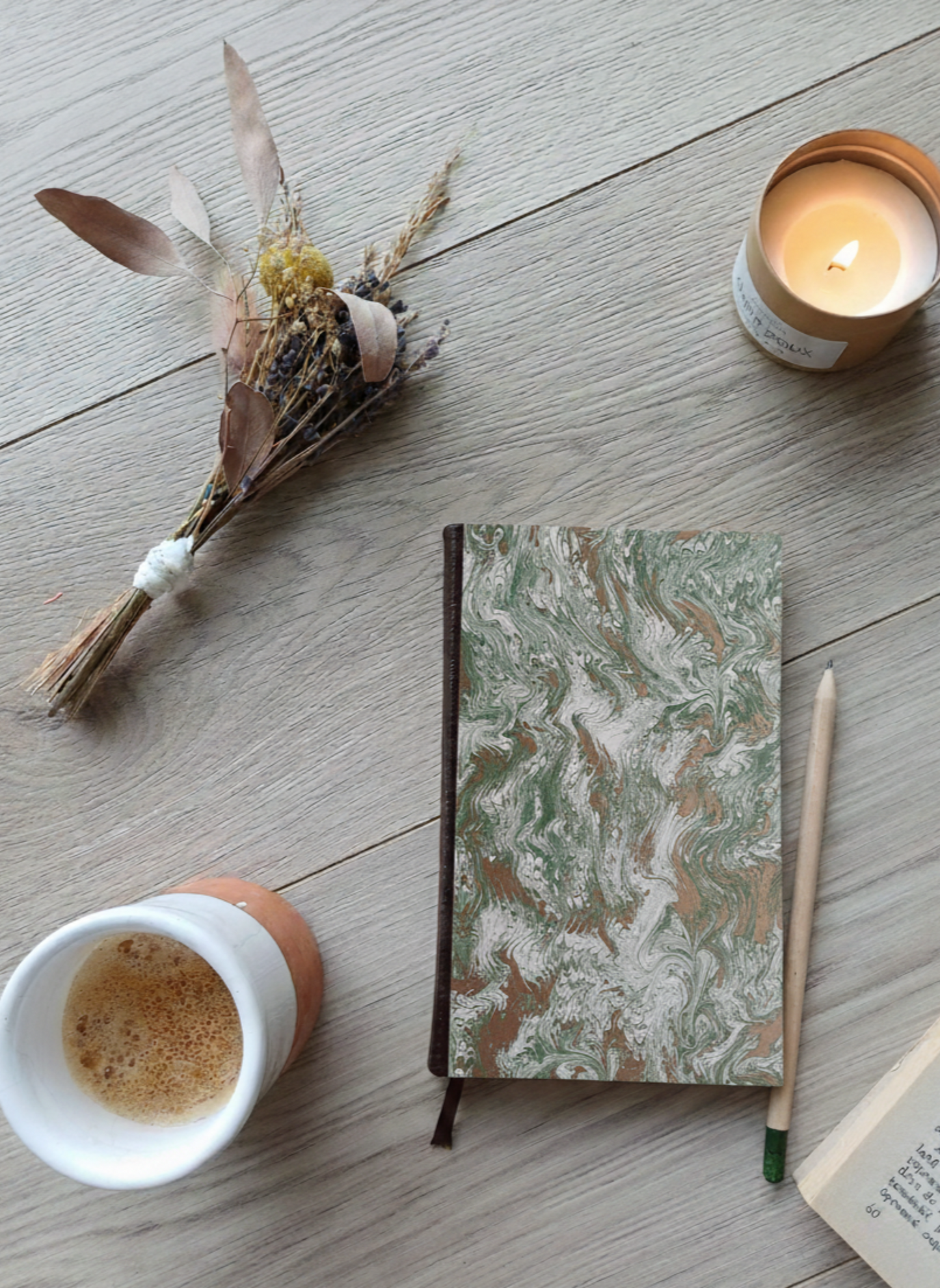 Carnet à la couverture imprimée avec des motifs marbrés verts et dorés, entouré de fleurs, d'un café, d'une bougie, d'un livre et d'un crayon.