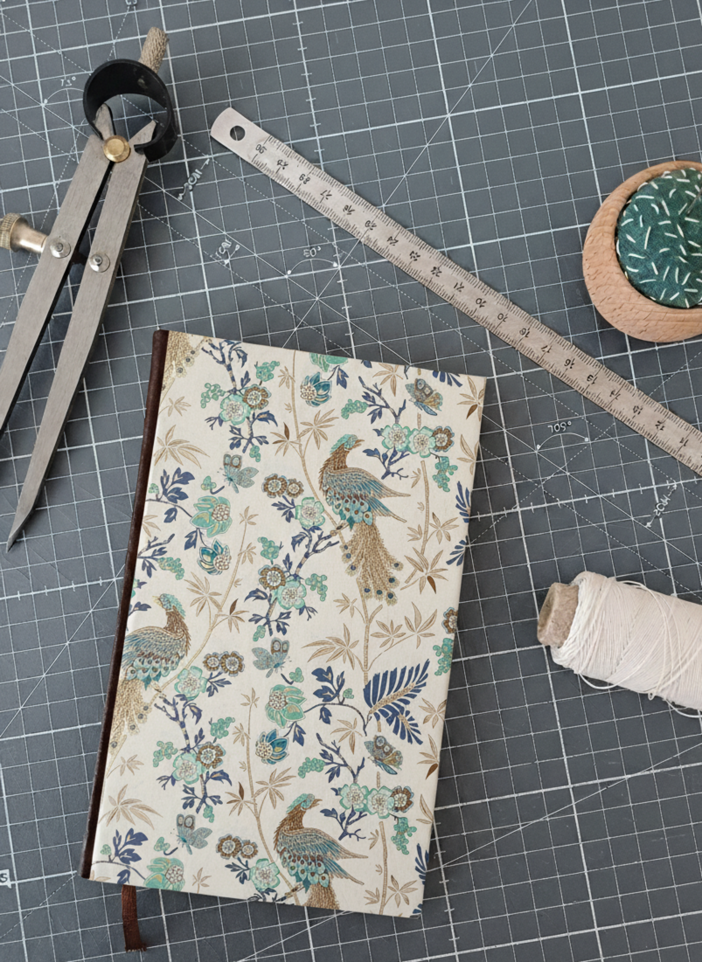 Carnet à la couverture imprimée de motifs fleuris et de paons dorés et bleus, entouré d'un compas, d'un réglet d'aiguilles et de fil.