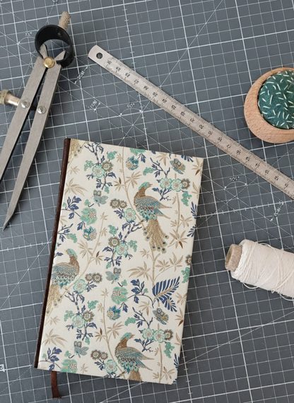 Carnet à la couverture imprimée de motifs fleuris et de paons dorés et bleus, entouré d'un compas, d'un réglet d'aiguilles et de fil.