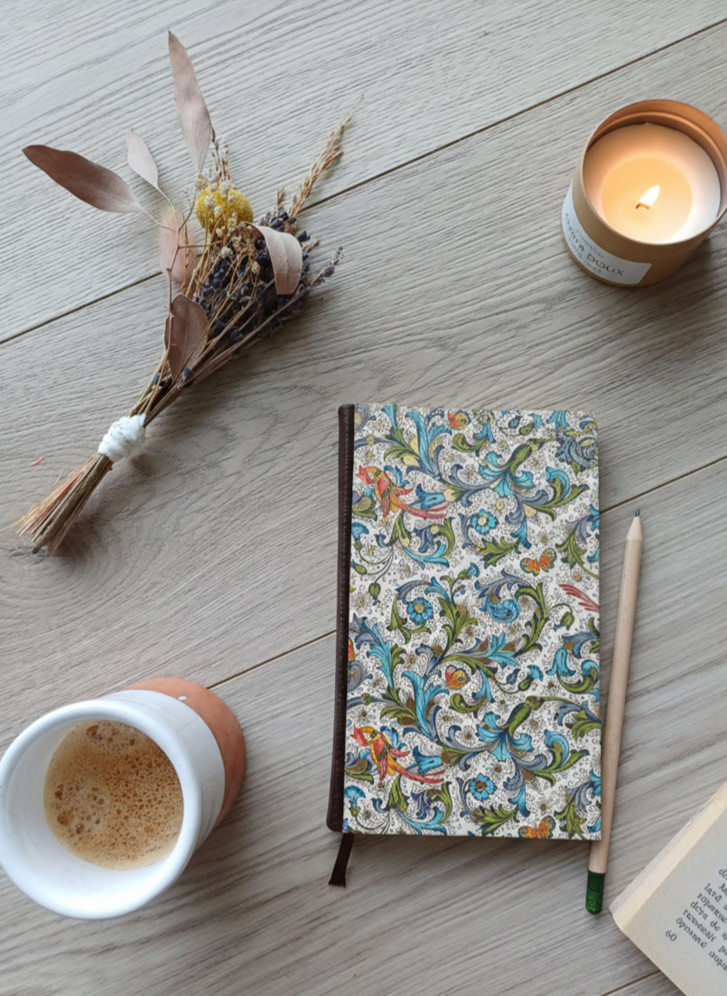 Carnet à la couverture imprimée avec des motifs fleuris et des paons, entouré de fleurs, d'un café, d'une bougie, d'un livre et d'un crayon.