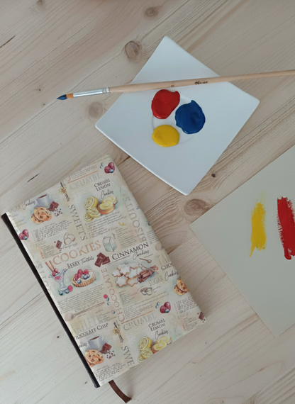 Carnet à la couverture imprimé de recettes et patisseries à côté d'une palette de peinture et un pinceau.