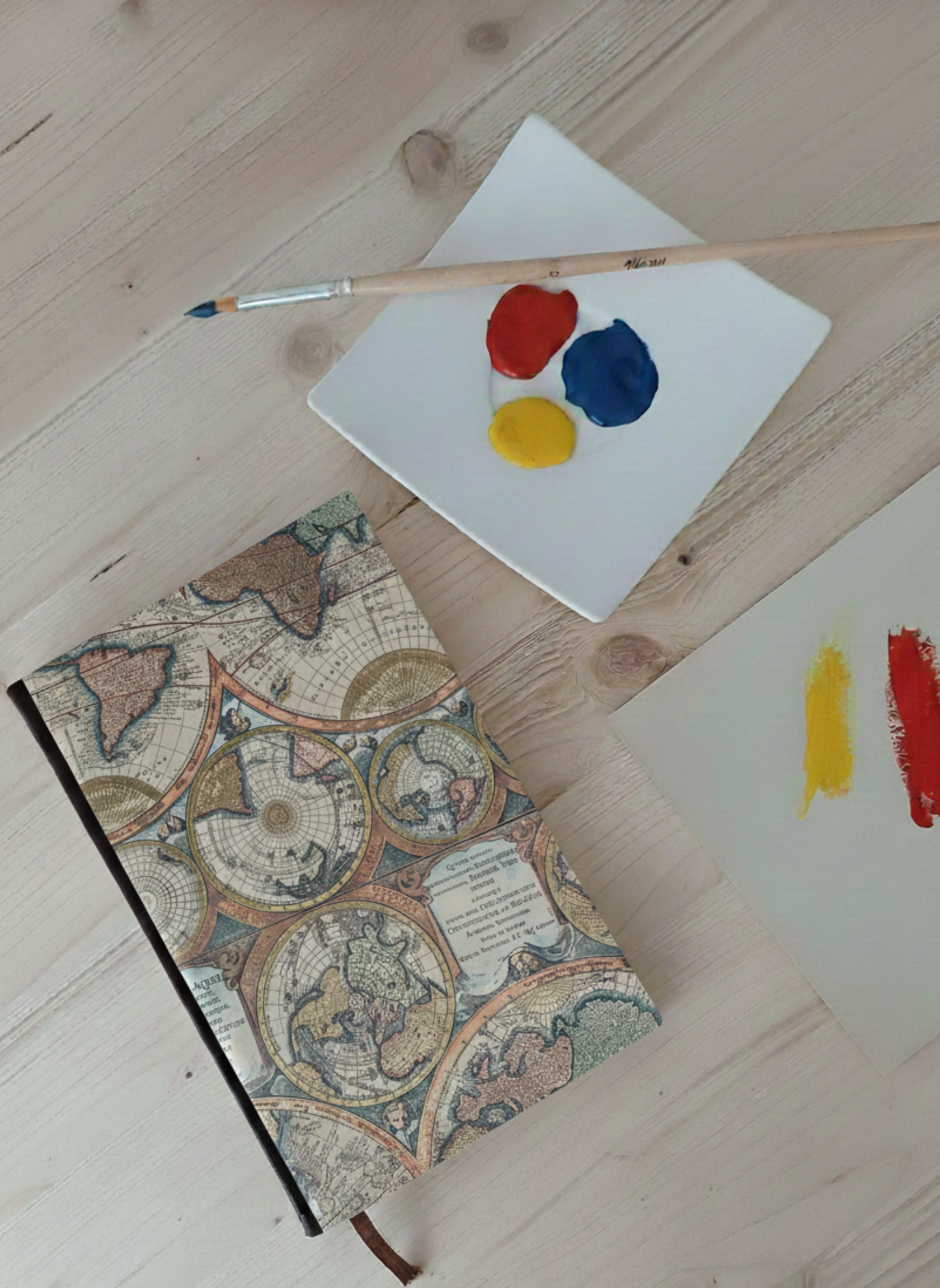 Carnet à la couverture imprimée de planisphères à côté d'une palette de peinture et un pinceau.