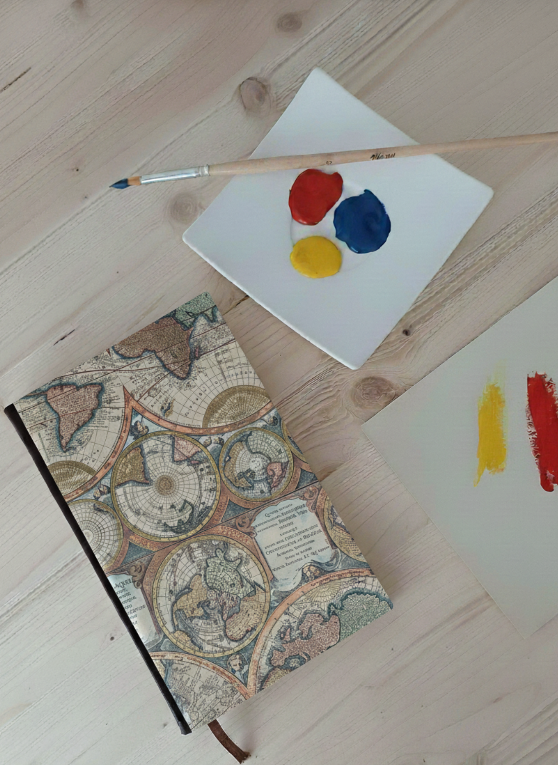 Carnet à la couverture imprimée de planisphères à côté d'une palette de peinture et un pinceau.
